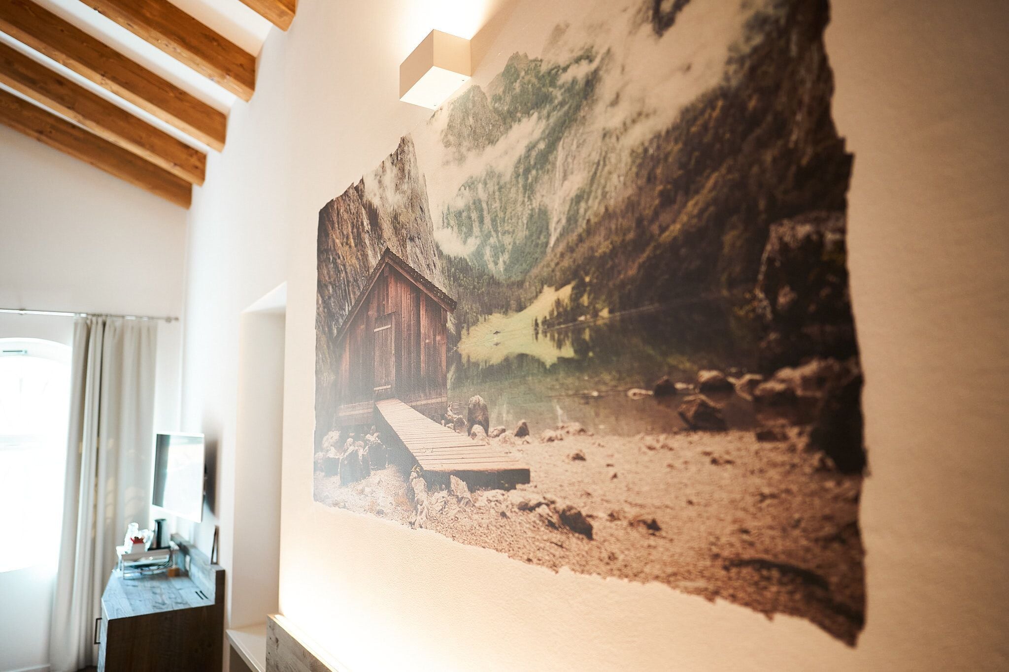 Una habitación con techo de madera, un cuadro colgado que muestra una cabaña junto a un lago, un pequeño escritorio de madera con una lámpara y una ventana con cortinas.