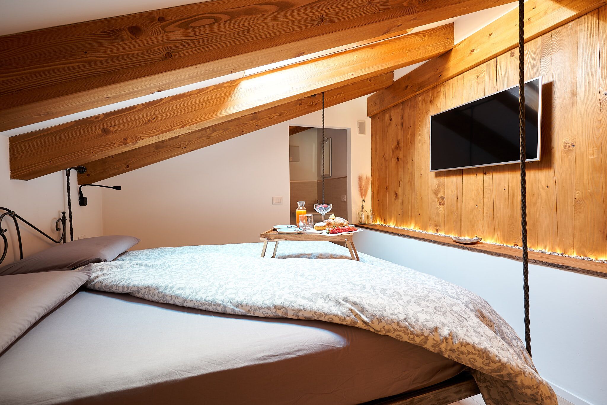 Ein Schlafzimmer mit schräg verlaufender Decke, Holzbalken, einem Bett mit gemusterten Bettbezügen, einem Nachttisch mit Saftglas und Speisenplatte sowie einem an der Wand befestigten Fernseher.