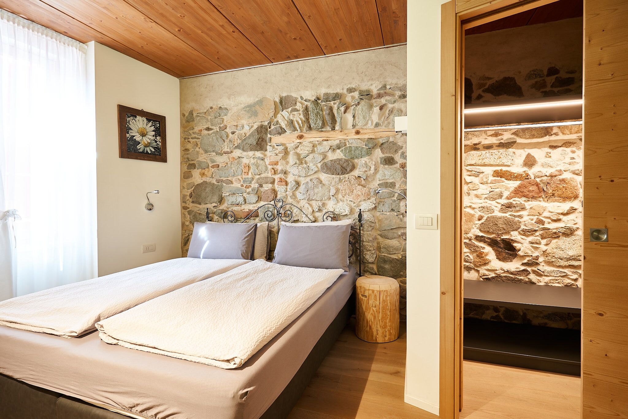 Ein Schlafzimmer mit einer Steinswand, einem Bett mit zwei Kissen, Holzboden und einem kleinen Holzschemel.