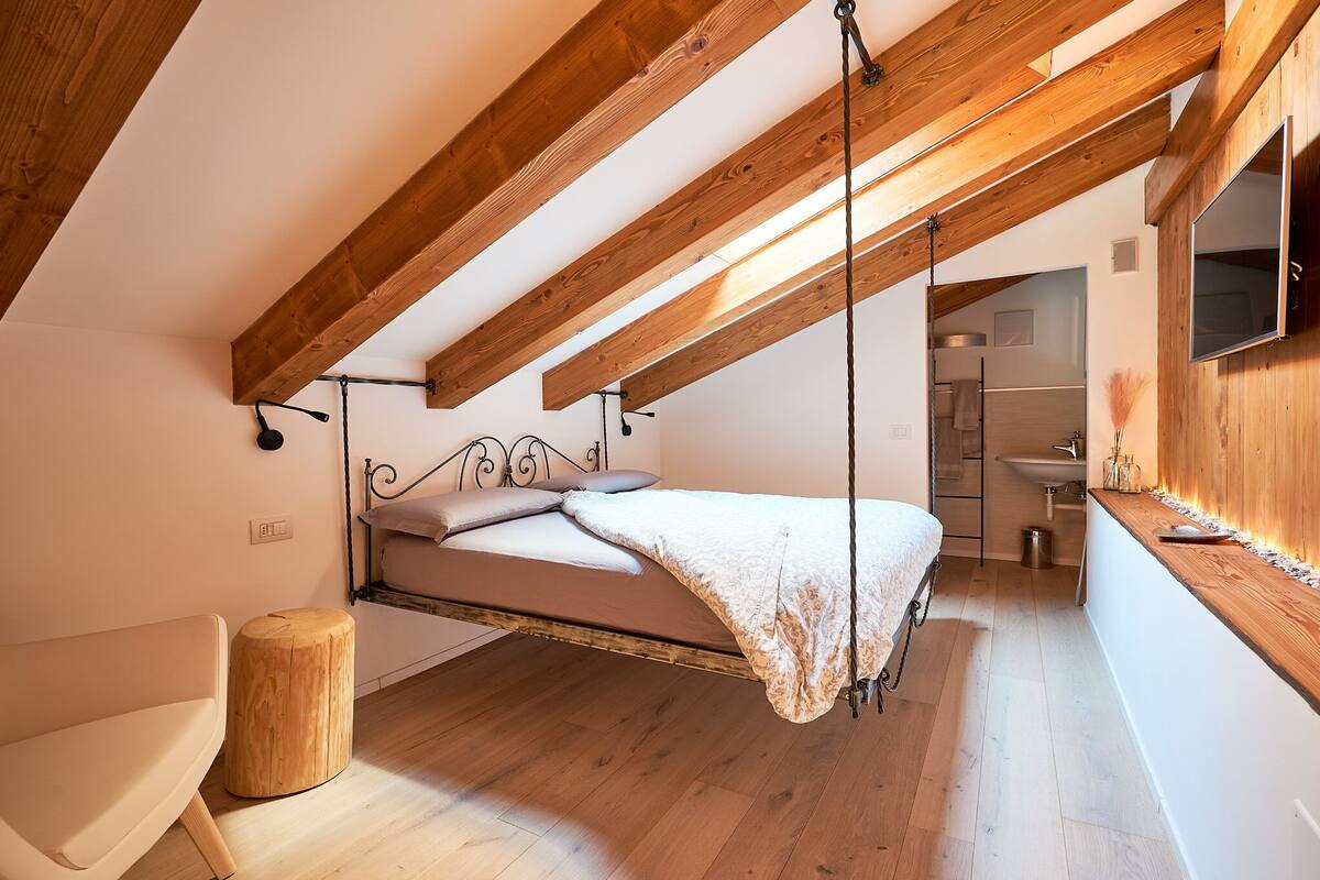 Ein Schlafzimmer im Loft-Stil mit einem von Holzbalken hängenden Bett, einem Holzschemel und Blick ins Bad.
