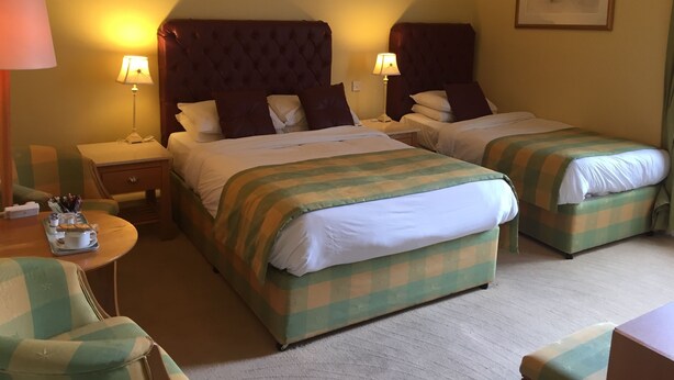 A hotel room with two beds, each with a checkered bedspread, a tufted headboard, and bedside lamps. There is a small table with a tray of breakfast items, a chair, and a framed picture on the wall. - Faythe Guesthouse (Wexford)