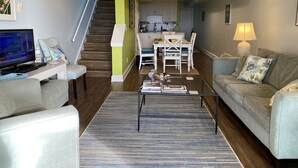 A living room with a sofa, coffee table, and TV, a staircase leading to a kitchen, and a dining area with chairs and a table. - OCEANFRONT Midisle Prvt Pool 3New Baths,Kitchen,Floors, AC,Total Renovation!     (Fripp Island)