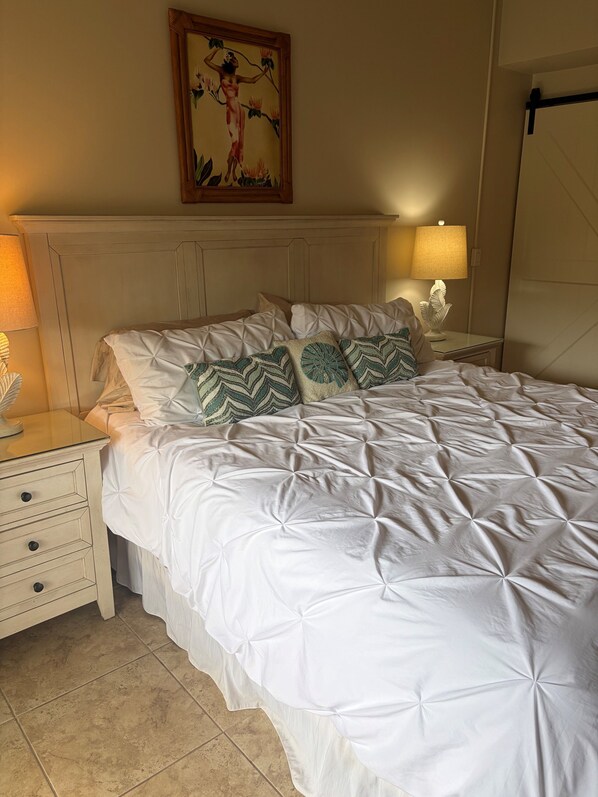 Una cama bien hecha con edredón blanco y almohadillas decorativas, cabecera de madera, mesita de noche con lámpara y una obra de arte enmarcada en la pared.