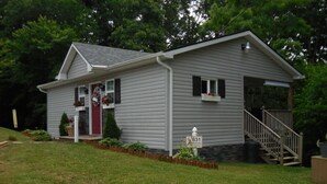 A single-story house with a red front door, a small porch with potted plants, a wooden staircase leading to a front door, and a mailbox.