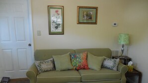 A green sofa with floral cushions, a framed floral painting, a green lamp, and a thermostat on the wall.