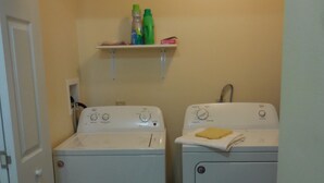 A laundry room with two white front-loading washing machines and a shelf above them holding cleaning supplies.