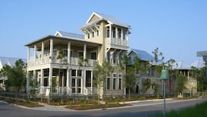 Une grande maison à deux étages, dotée d’un balcon au deuxième étage, entourée d’une pelouse bien entretenue et bordée d’une route au premier plan.