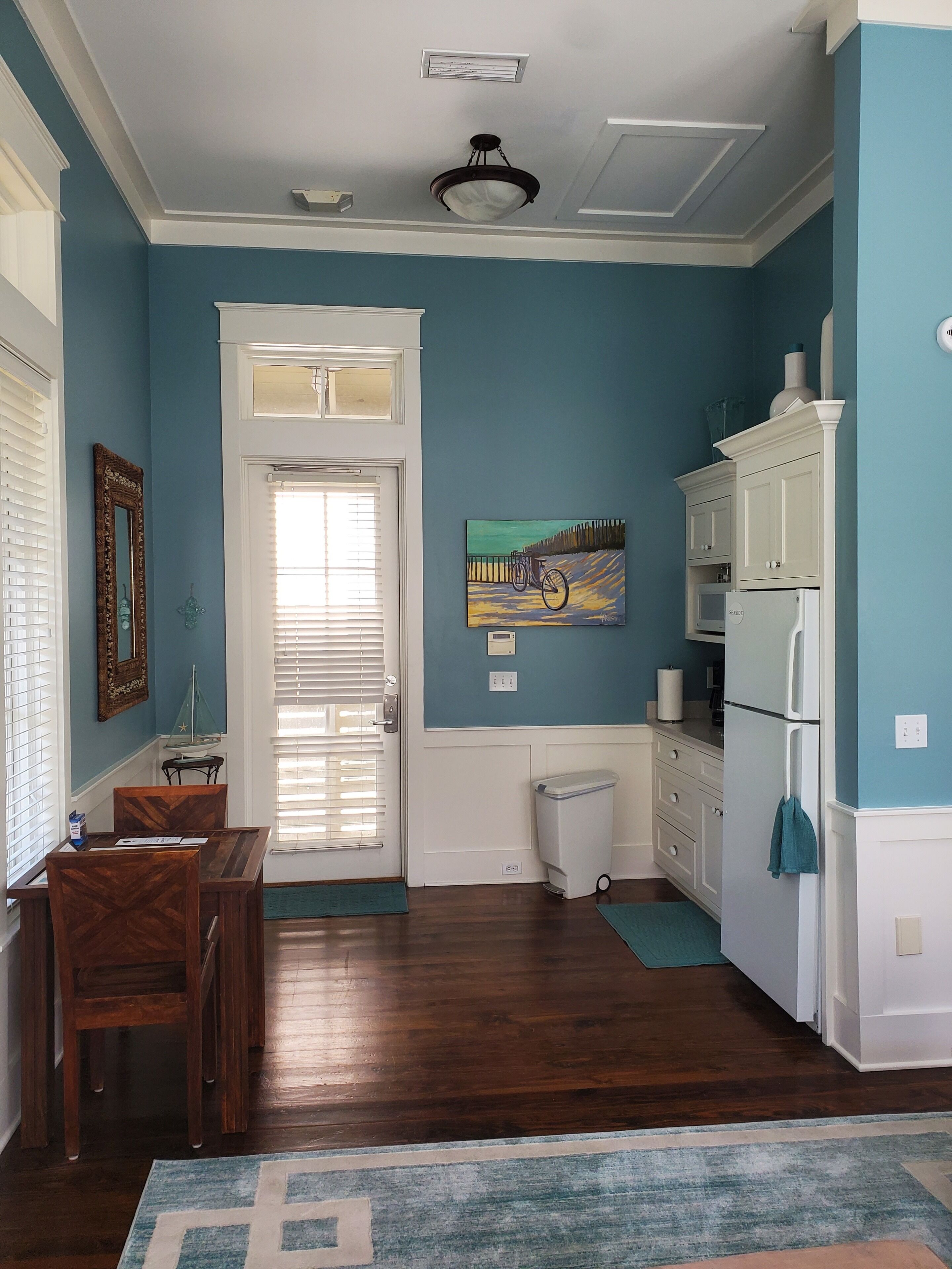 A kitchen with blue walls, white cabinetry, a dining table with chairs, a trash can, and a painting on the wall.