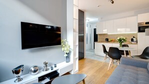 A modern living room with a flat-screen TV, a coffee table, and a sofa. There is a dining area with a table and chairs, and a kitchen in the background.