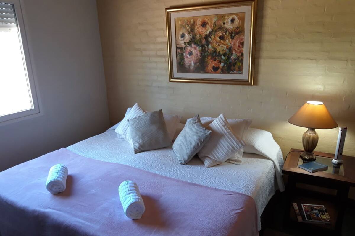 A neatly made bed with white linens and pillows, a framed floral painting on the wall, and a bedside lamp.