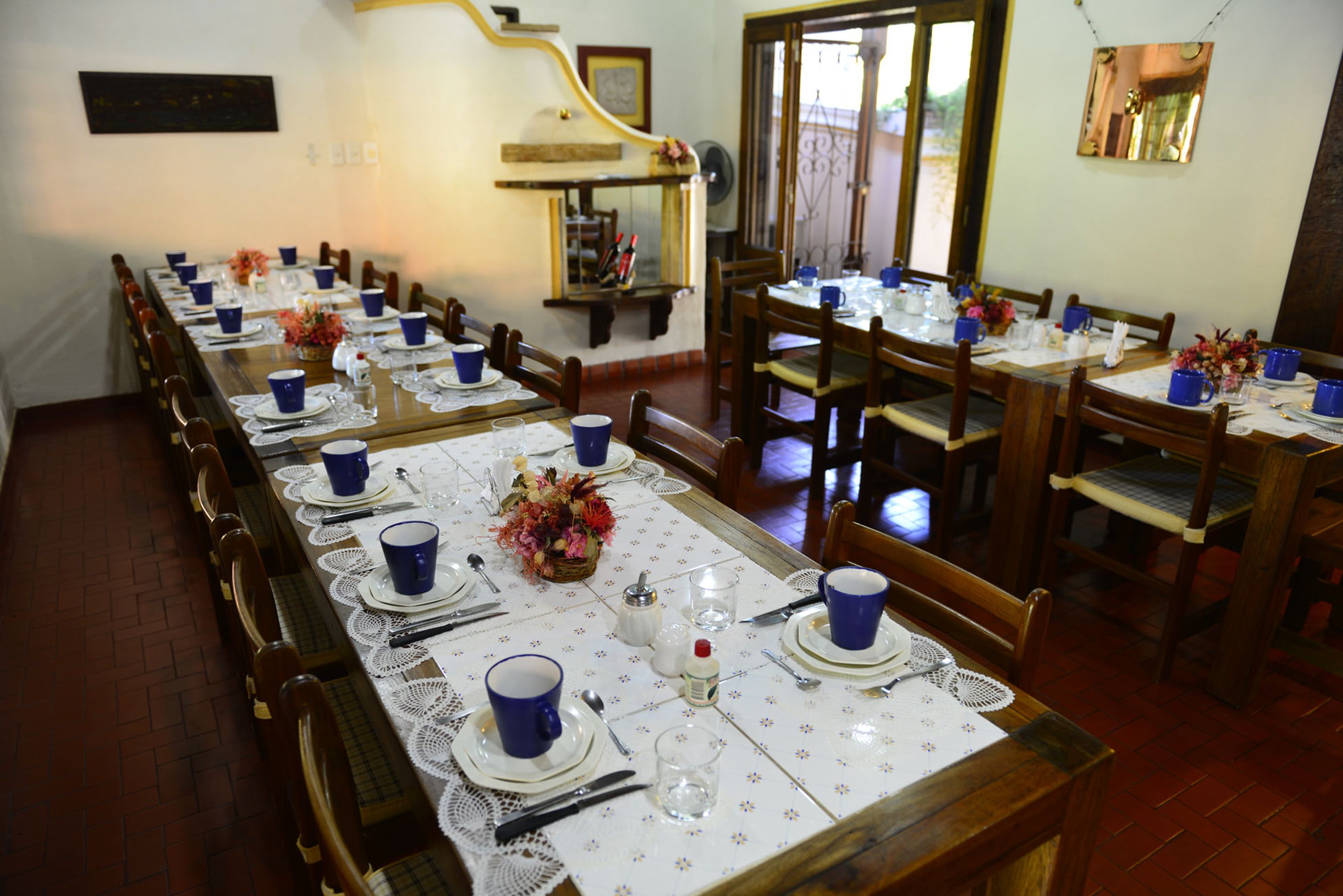 A dining area with long tables set for multiple people, featuring blue cups, white plates, and lace tablecloths.
