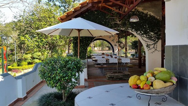 An outdoor dining area with tables and umbrellas, surrounded by greenery and a stone wall. - Hotel La Quinta (Paraguari)