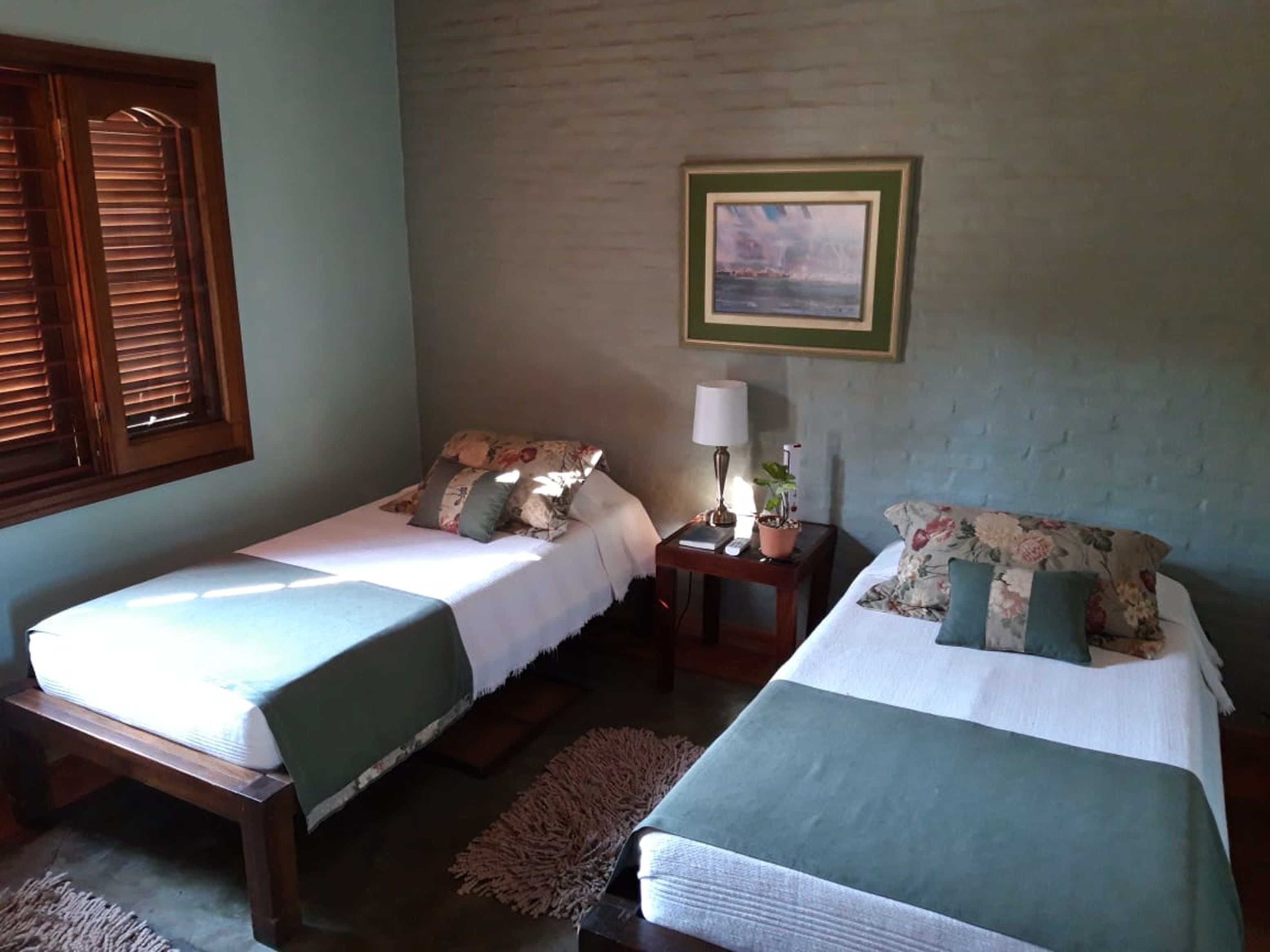 Una habitación con dos camas individuales, una mesita de noche con una lámpara y una planta, un cuadro en la pared y una ventana con contraventanas de madera.