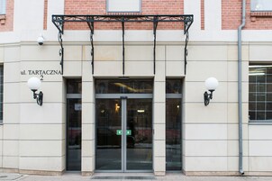 A building entrance with a glass door, a sign reading "ul. TARTACZNA 2", and two wall-mounted light fixtures.