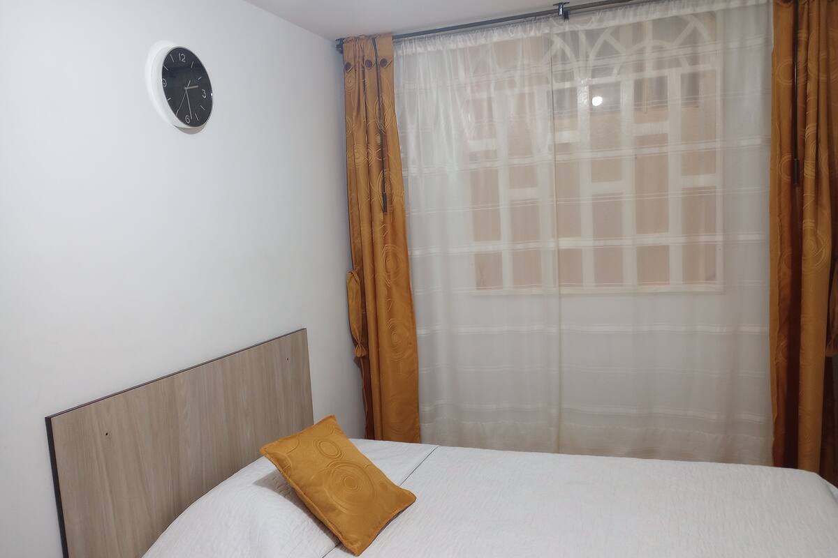 Une chambre à coucher avec un lit, une tête de lit, une horloge murale et une fenêtre avec des rideaux.