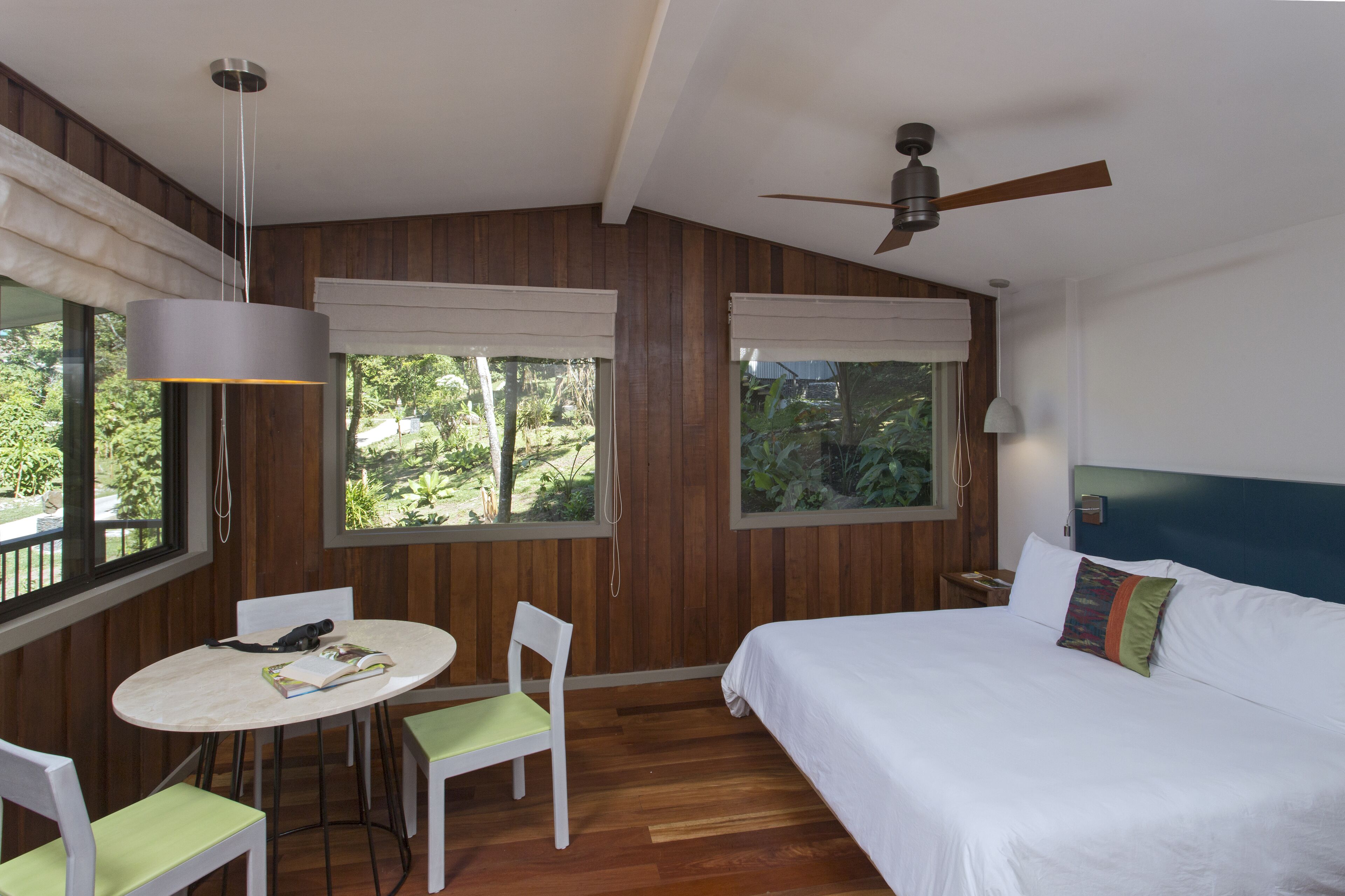 A bedroom with a bed, a ceiling fan, a table with chairs, and large windows with curtains.