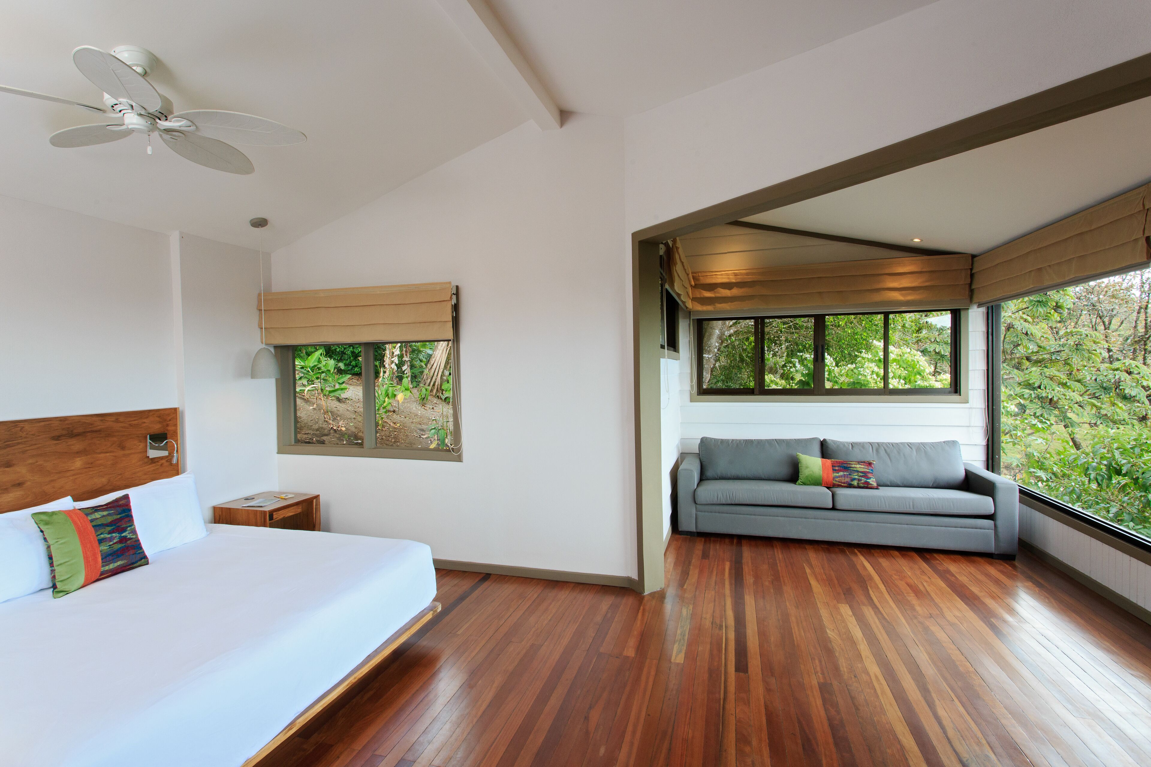 A bedroom with a bed, a gray sofa, a ceiling fan, and large windows with a view of greenery.