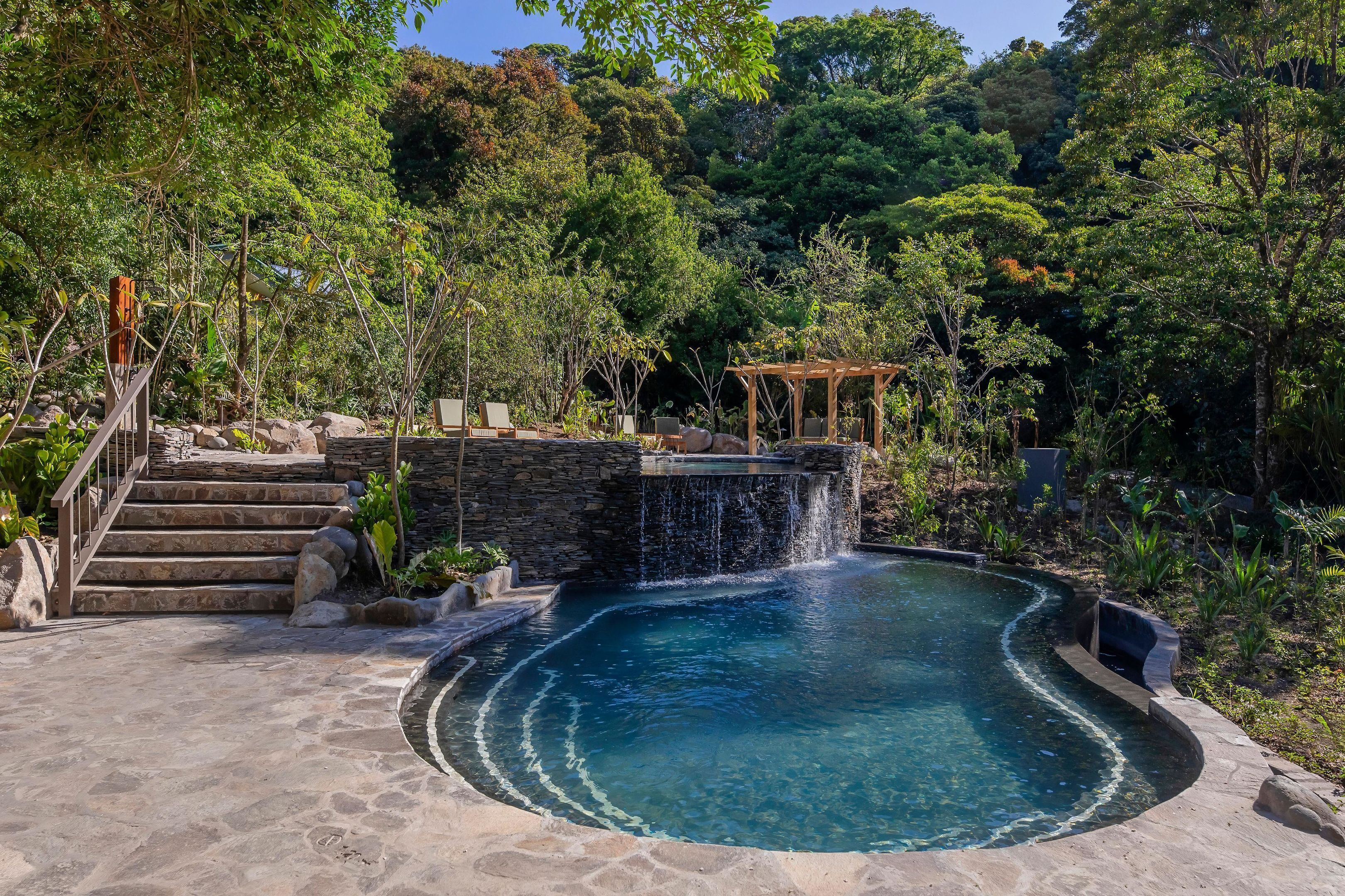 A circular pool with a waterfall feature, stone steps leading up to a wooden pergola, and lush greenery surrounding the area.