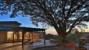 Un espace extérieur dallé de pierres, agrémenté d’une pergola, d’un grand arbre et d’un bâtiment doté de grandes fenêtres.