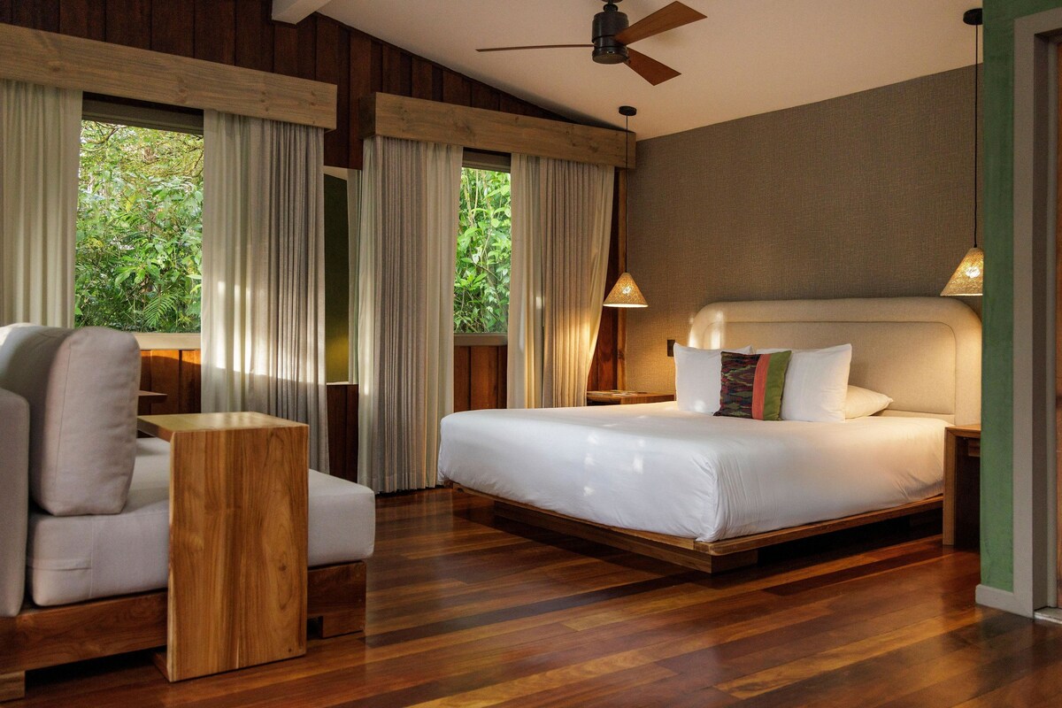 A bedroom with a bed, a wooden side table, a chair, and a ceiling fan.
