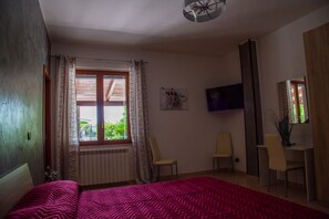 Un dormitorio con cama, ventana con cortinas, una silla, escritorio y televisor en la pared.