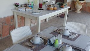 Un comedor con una mesa blanca dispuesta para cuatro personas, con platos, cubiertos y una tetera. Se aprecia una mesa de buffet con diversos alimentos y una planta en maceta al fondo.
