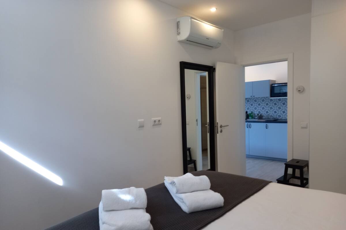 Quarto de hotel com uma cama, duas toalhas, um banquinho, um aparelho de ar condicionado e vista para a cozinha por uma porta aberta.