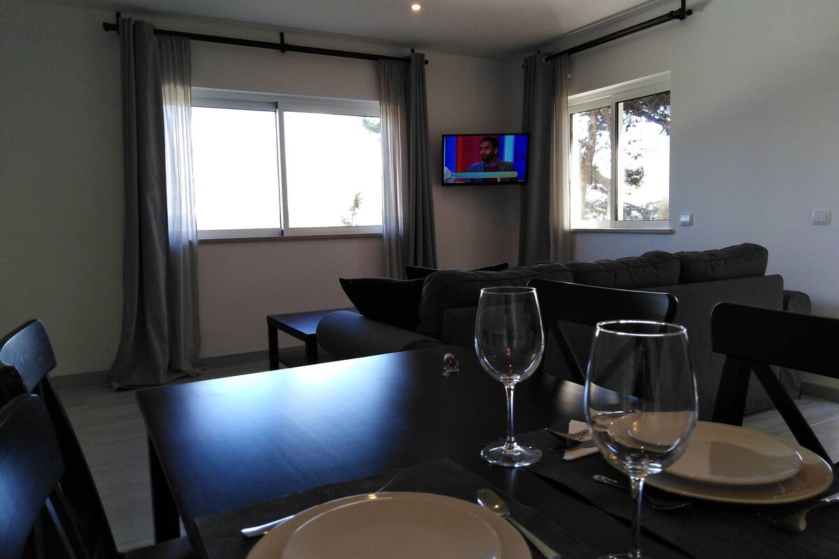 Uma sala de jantar com mesa posta para dois, uma TV na parede e um sofá ao fundo.