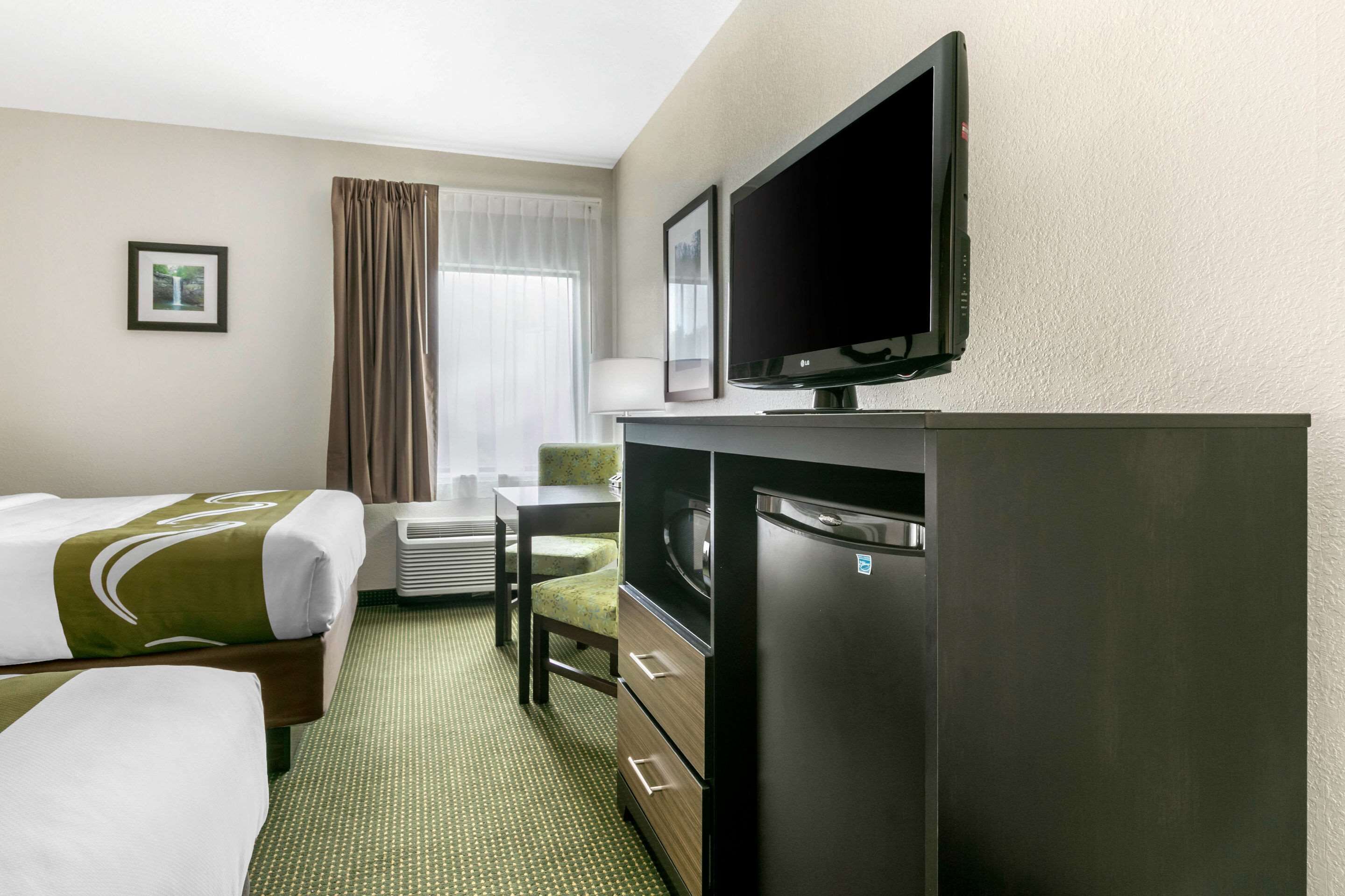 A hotel room with two beds, a flat-screen TV, a refrigerator, a microwave, a dining table with a chair, and a window with curtains.