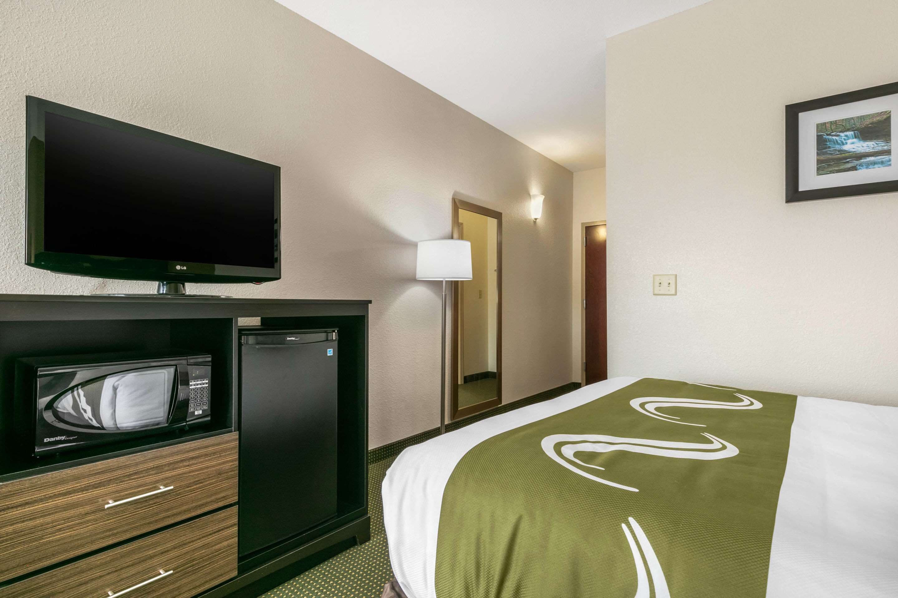 A hotel room with a flat-screen TV, a microwave, a mini-fridge, a bed with a green and white blanket, and a framed picture on the wall.