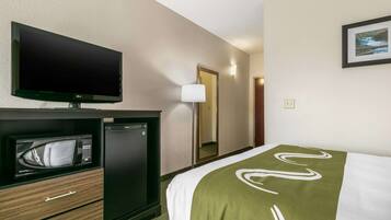 A hotel room with a flat-screen TV, a microwave, a mini-fridge, a bed with a green and white blanket, and a framed picture on the wall.