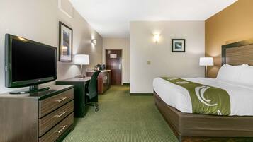 Quarto de hotel com uma cama, uma escrivaninha com computador, uma televisão e uma porta que dá para outro quarto.