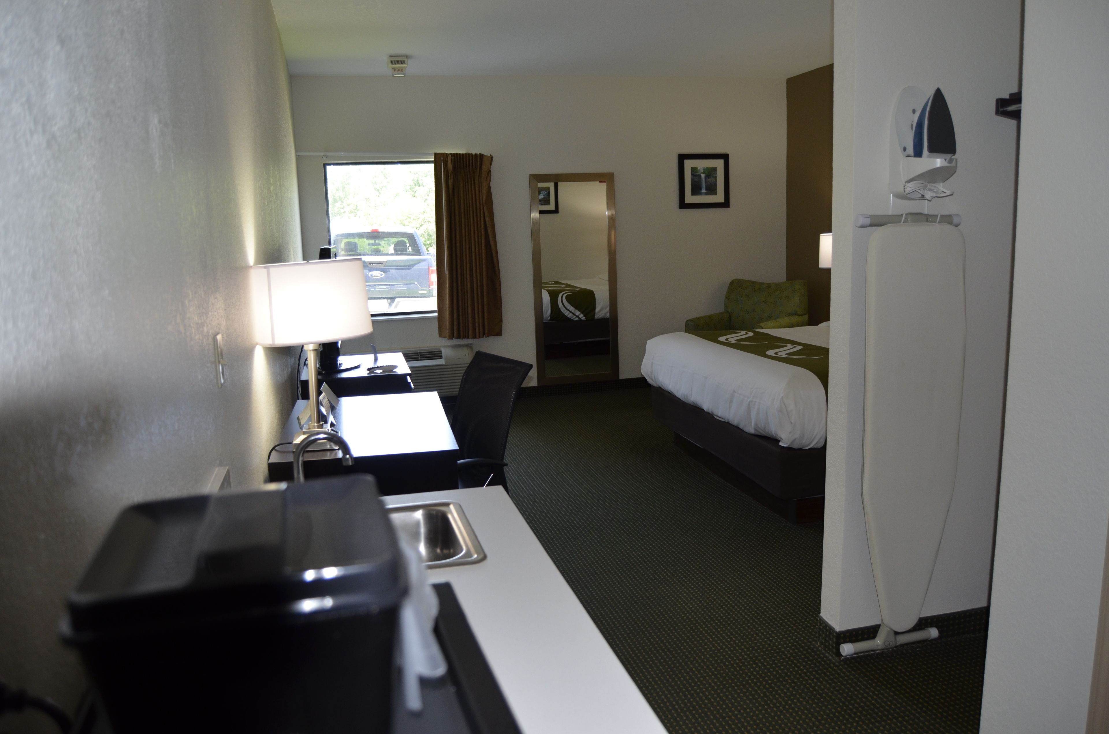 Habitación de hotel con una cama, un escritorio con lámpara, una silla, un espejo y una ventana con cortinas.