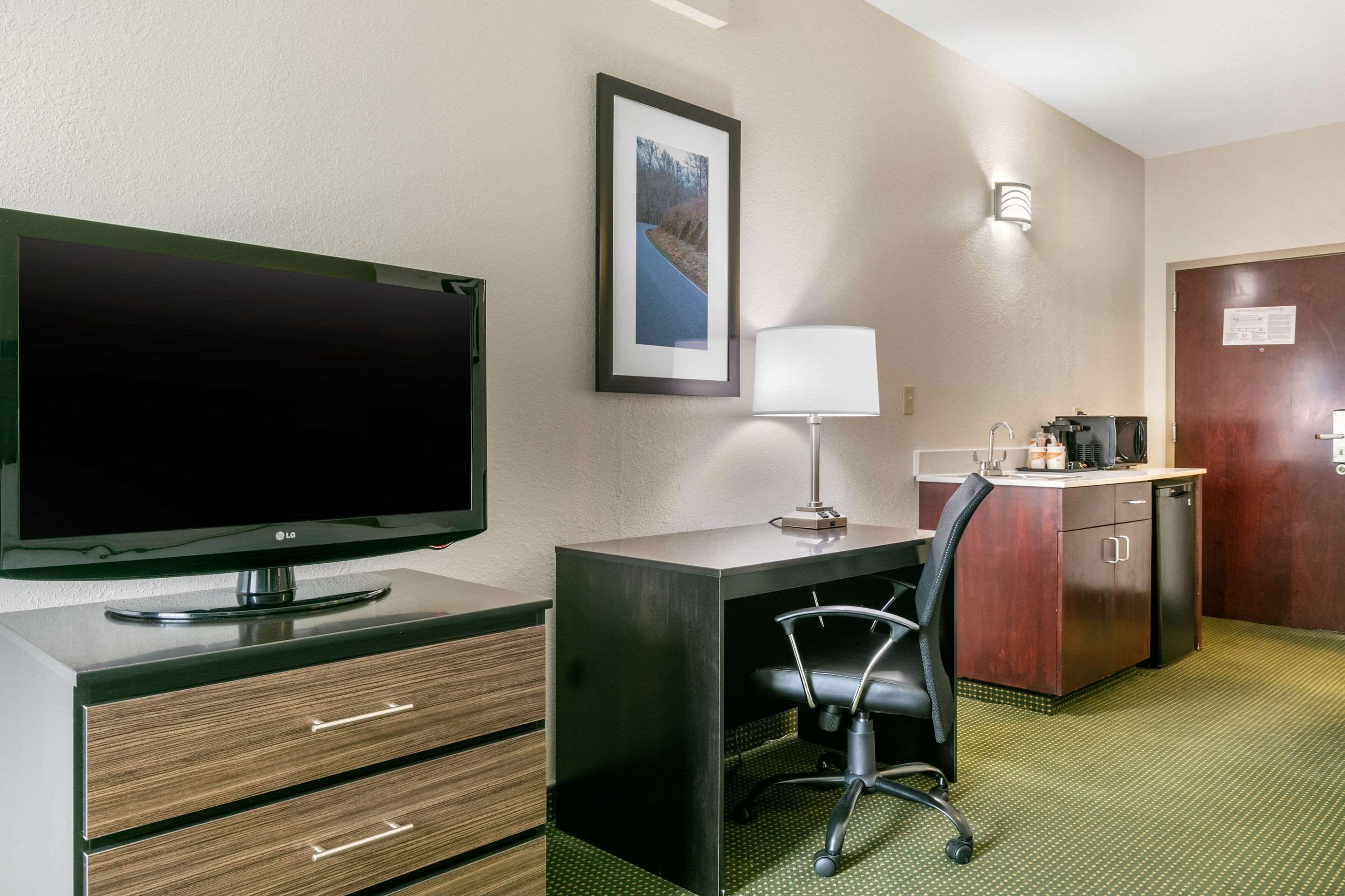 A hotel room with a desk, chair, TV, lamp, and a small kitchenette.