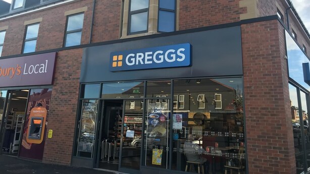 Ein Gebäude mit einem Greggs-Geschäft im Erdgeschoss, einem Sainsbury's Local darüber und einem sichtbaren Geldautomaten links.
