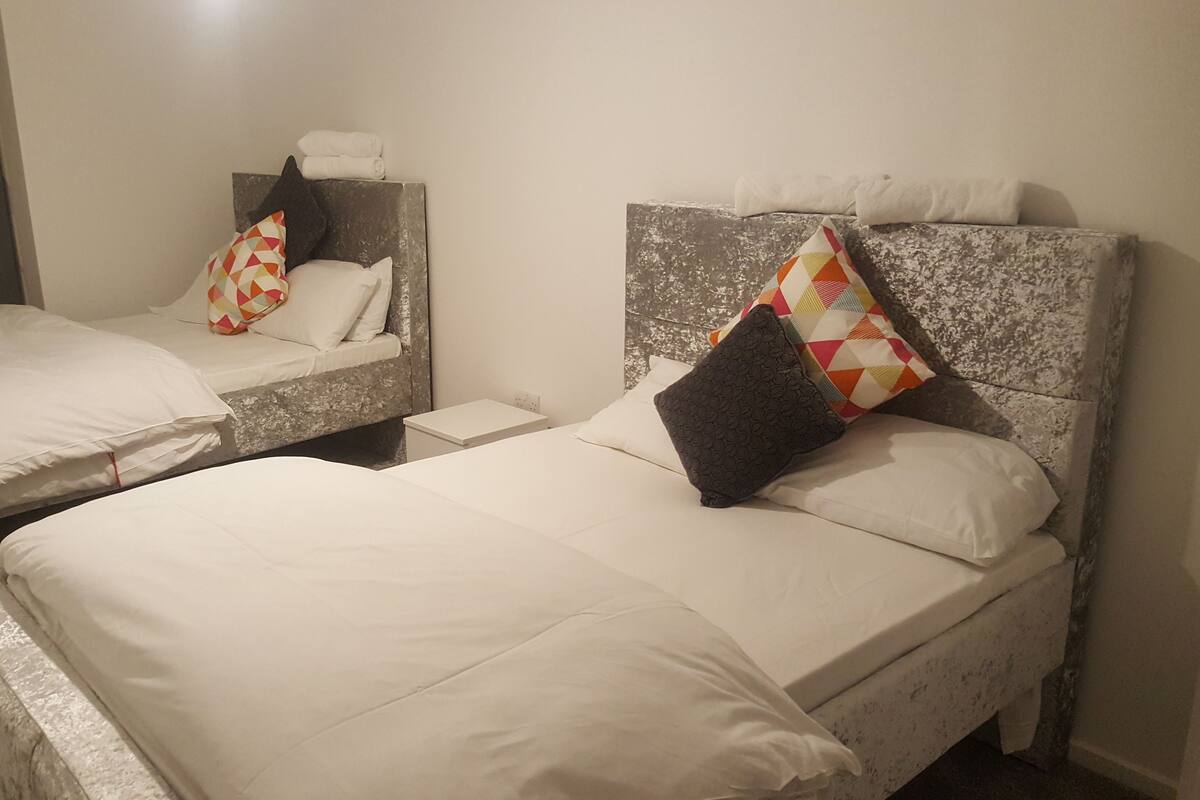 Two beds with white linens, a headboard with a geometric patterned pillow, and a small bedside table.