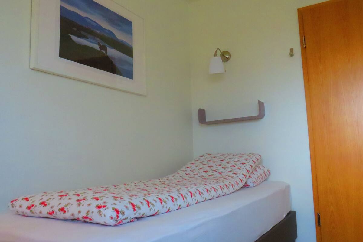 Ein kleines Zimmer mit einem Einzelbett, einem blumengemusterten Matratze, einer an der Wand befestigten Nachttischlampe und einem gerahmten Bild über dem Bett.