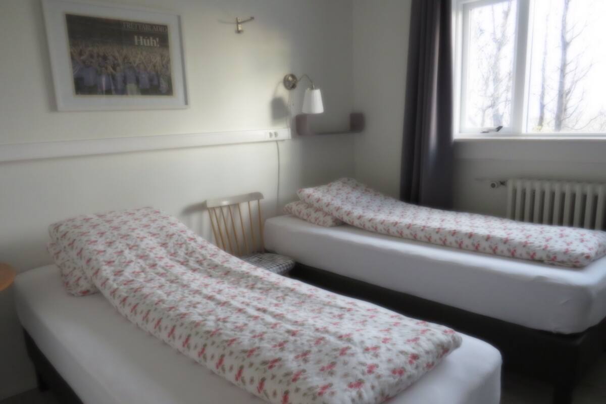 Ein Zimmer mit zwei Einzelbetten, einem kleinen Tisch, einem Stuhl, einem Heizkörper, einem gerahmten Bild an der Wand und einem Fenster mit Blick auf Bäume.