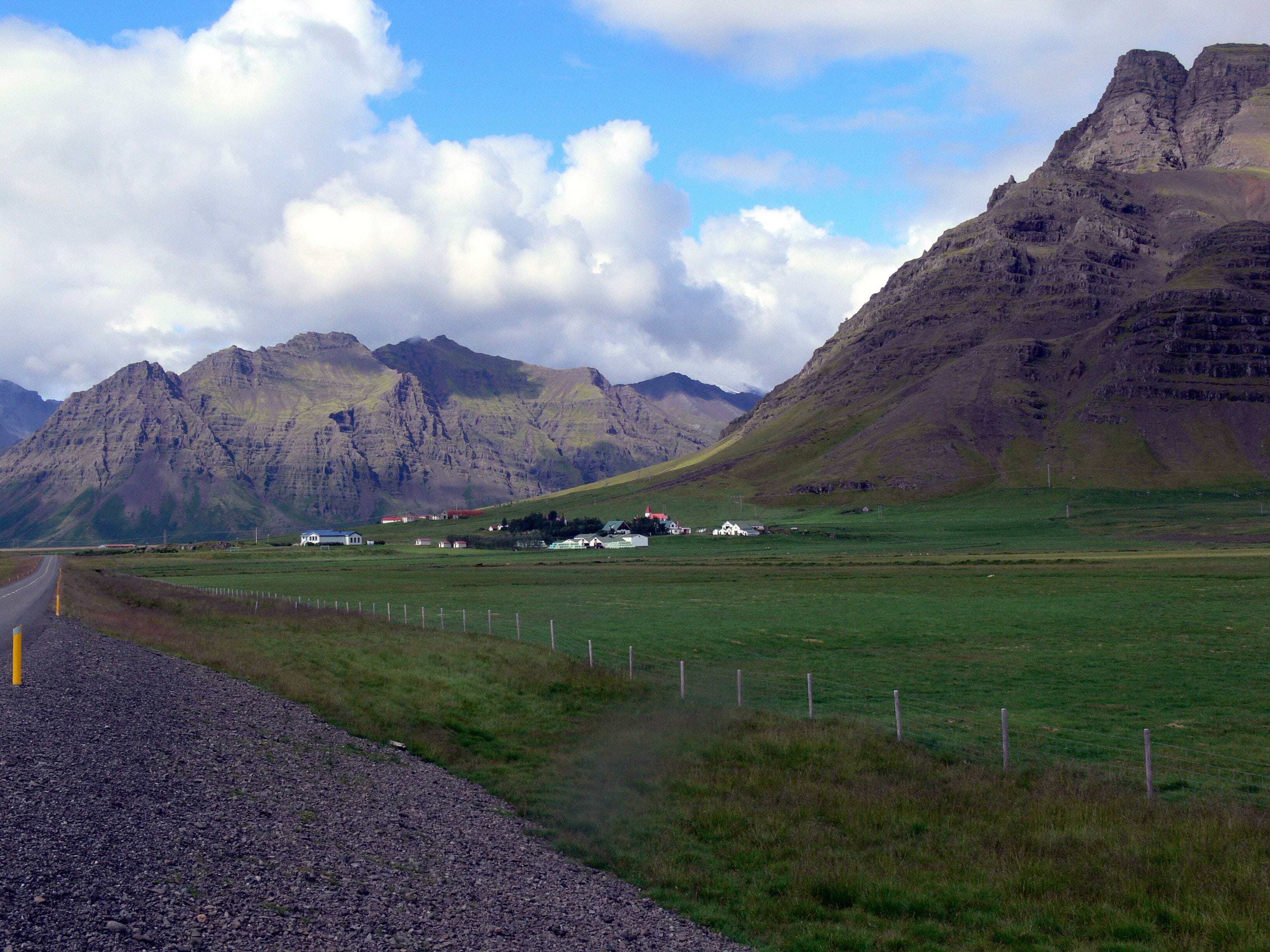 A rural landscape with a road, grassy fields, and a cluster of houses with a mountain backdrop.
