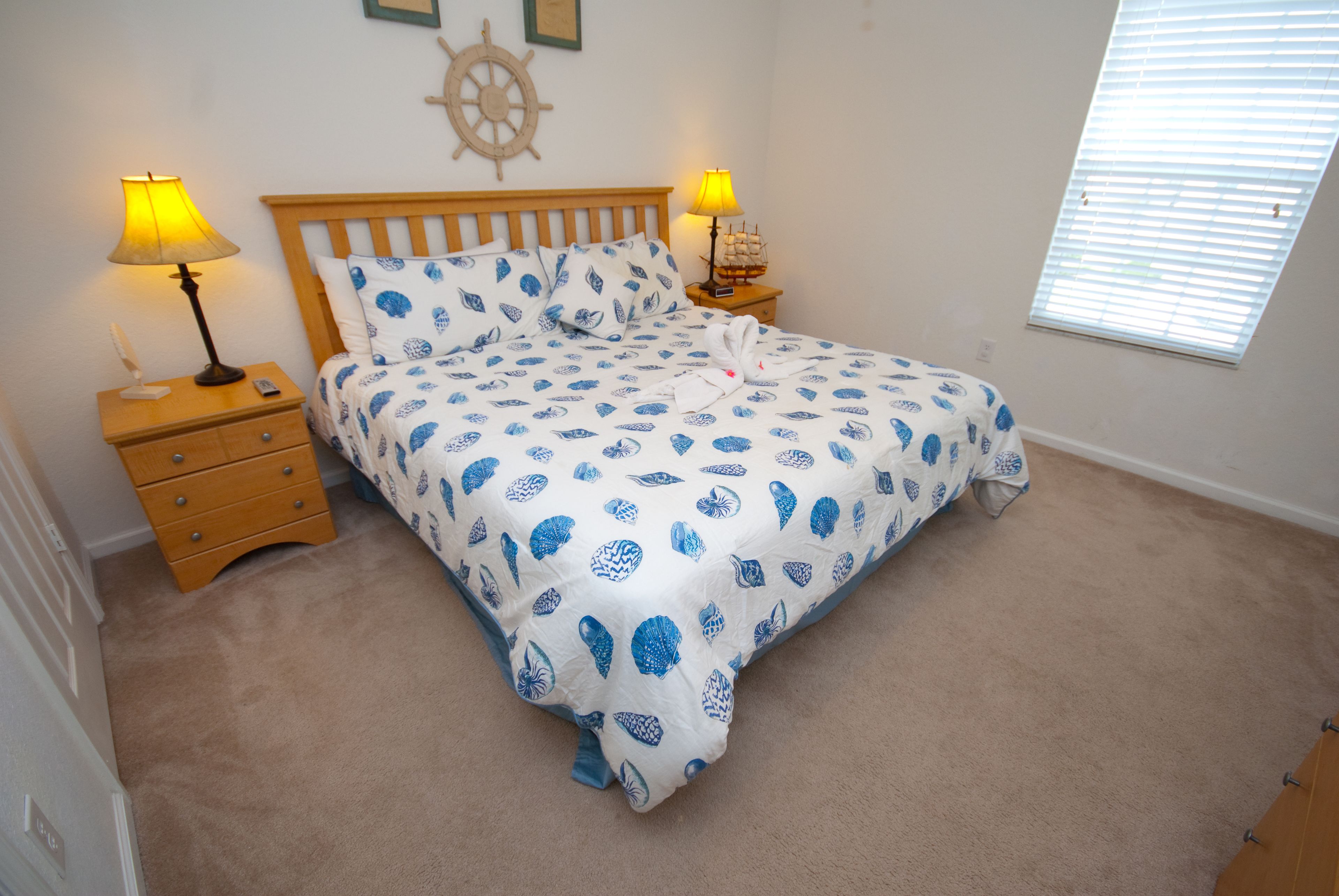 A neatly made bed with a patterned bedspread, two bedside lamps, a wooden nightstand with drawers, a ship's wheel decoration on the wall, and a window with blinds.