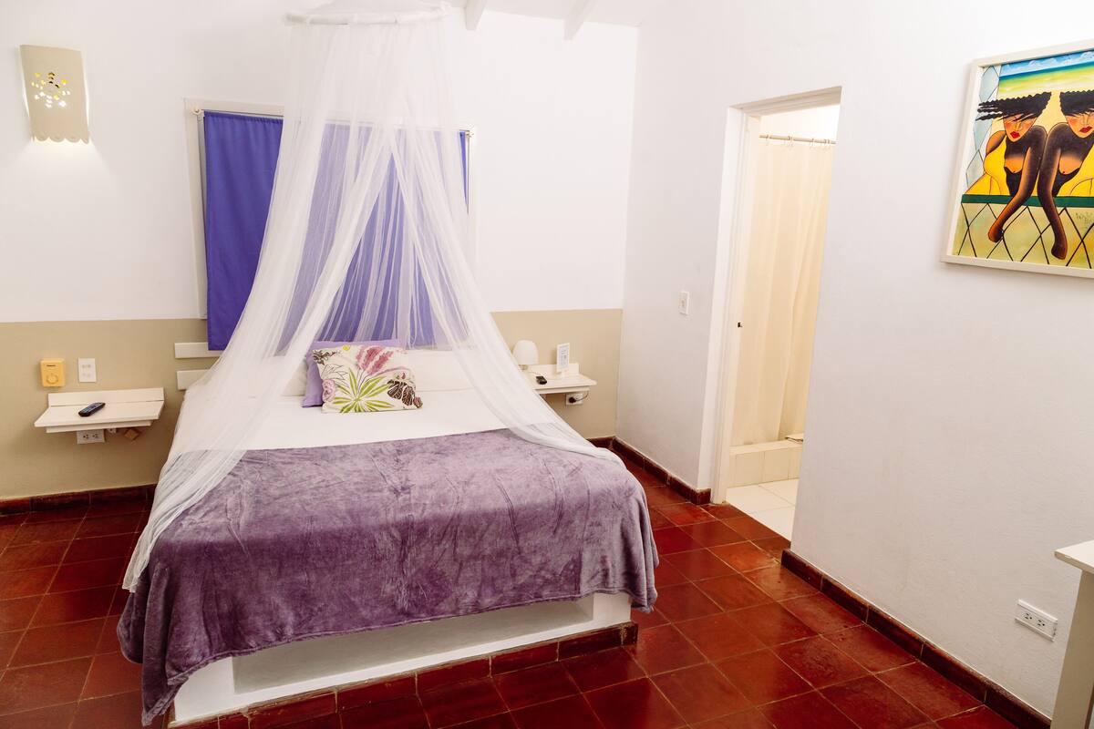 A bedroom with a bed, a nightstand, a bedside table, a wall-mounted light, a painting, and a door.
