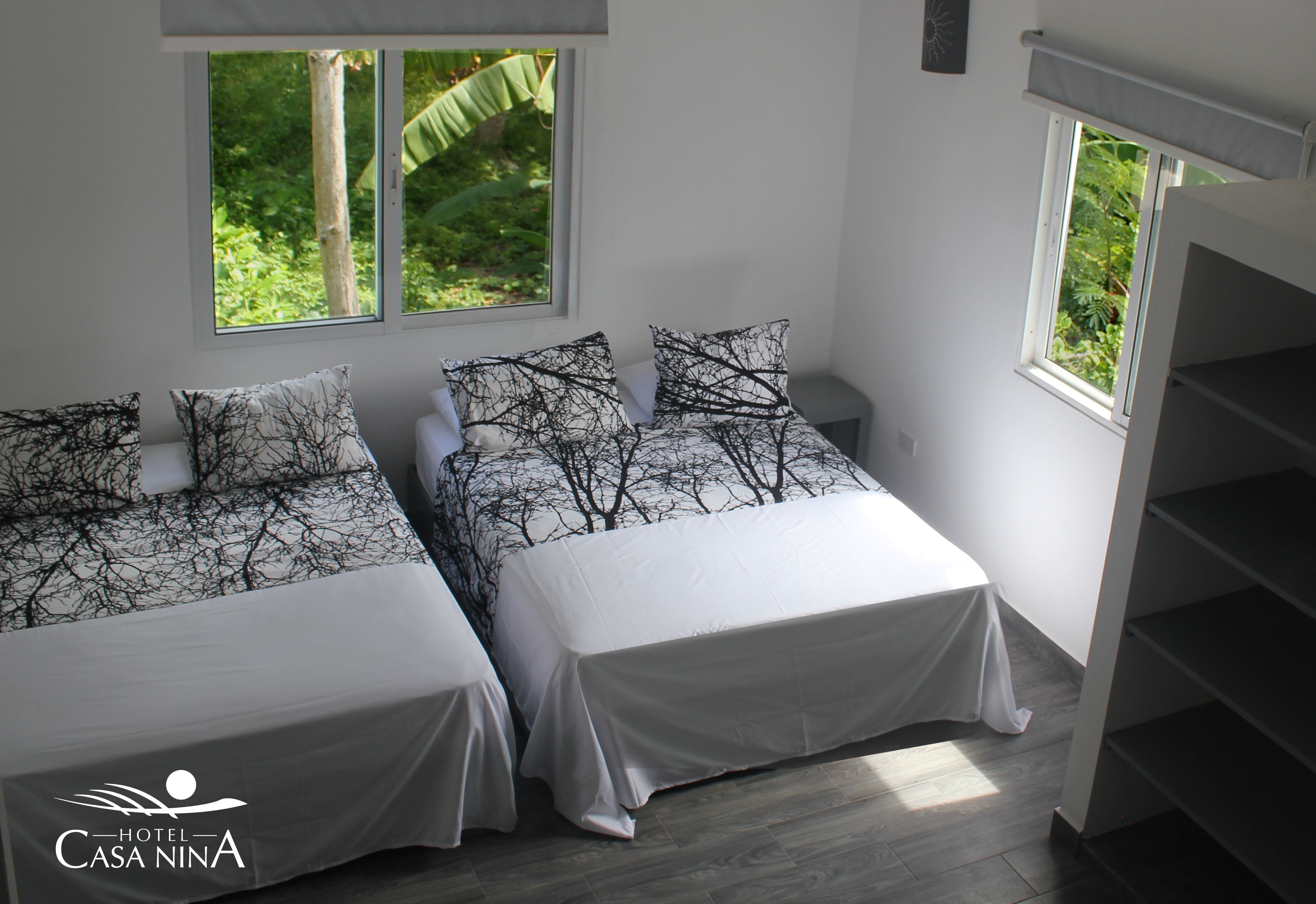 A hotel room with two beds, each with a white bedspread and black tree-patterned pillows. There are windows providing views of greenery outside.