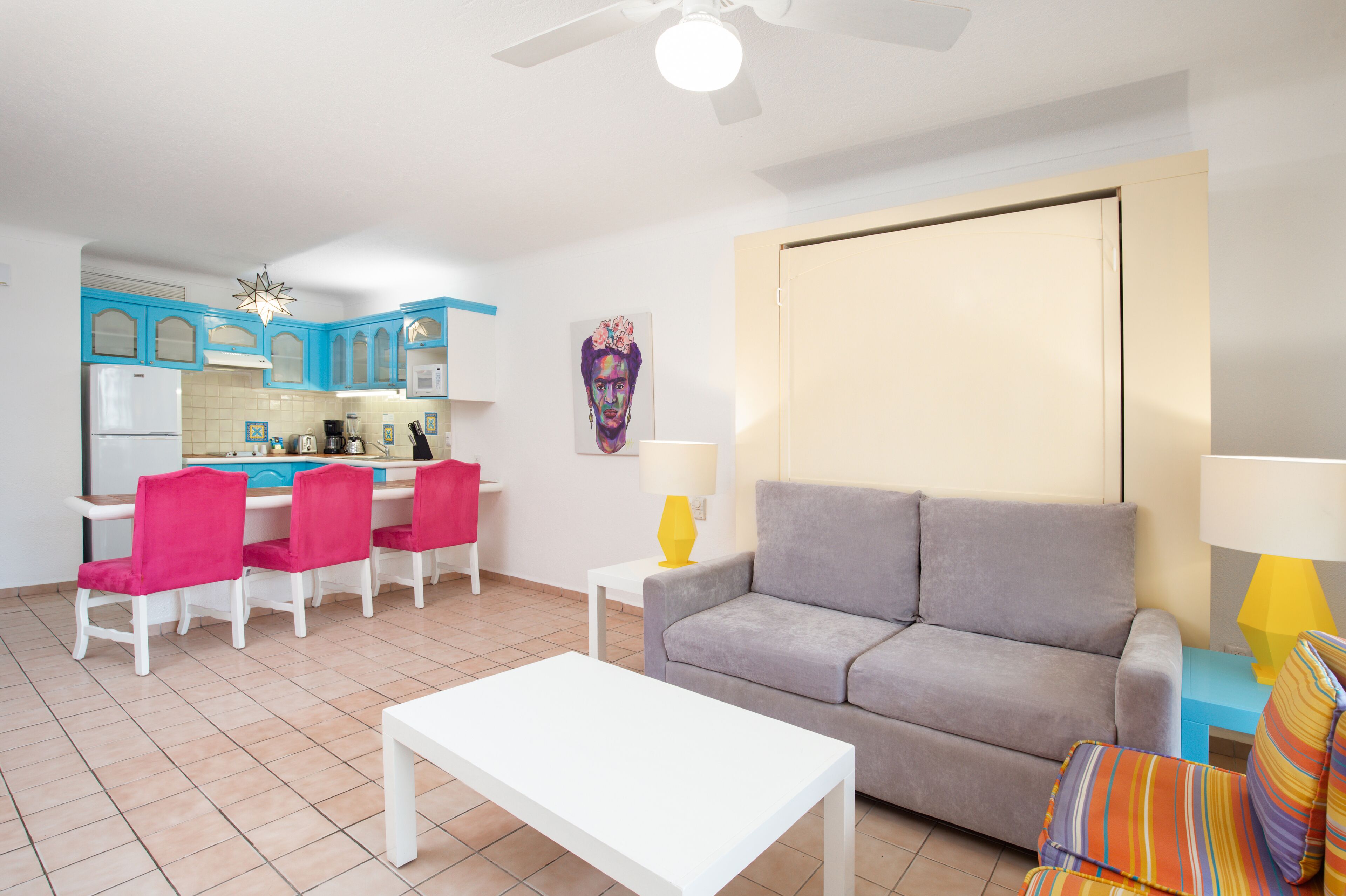 Una sala de estar con un sofá gris, una mesa de centro blanca y un comedor con sillas rosadas y armarios azules.