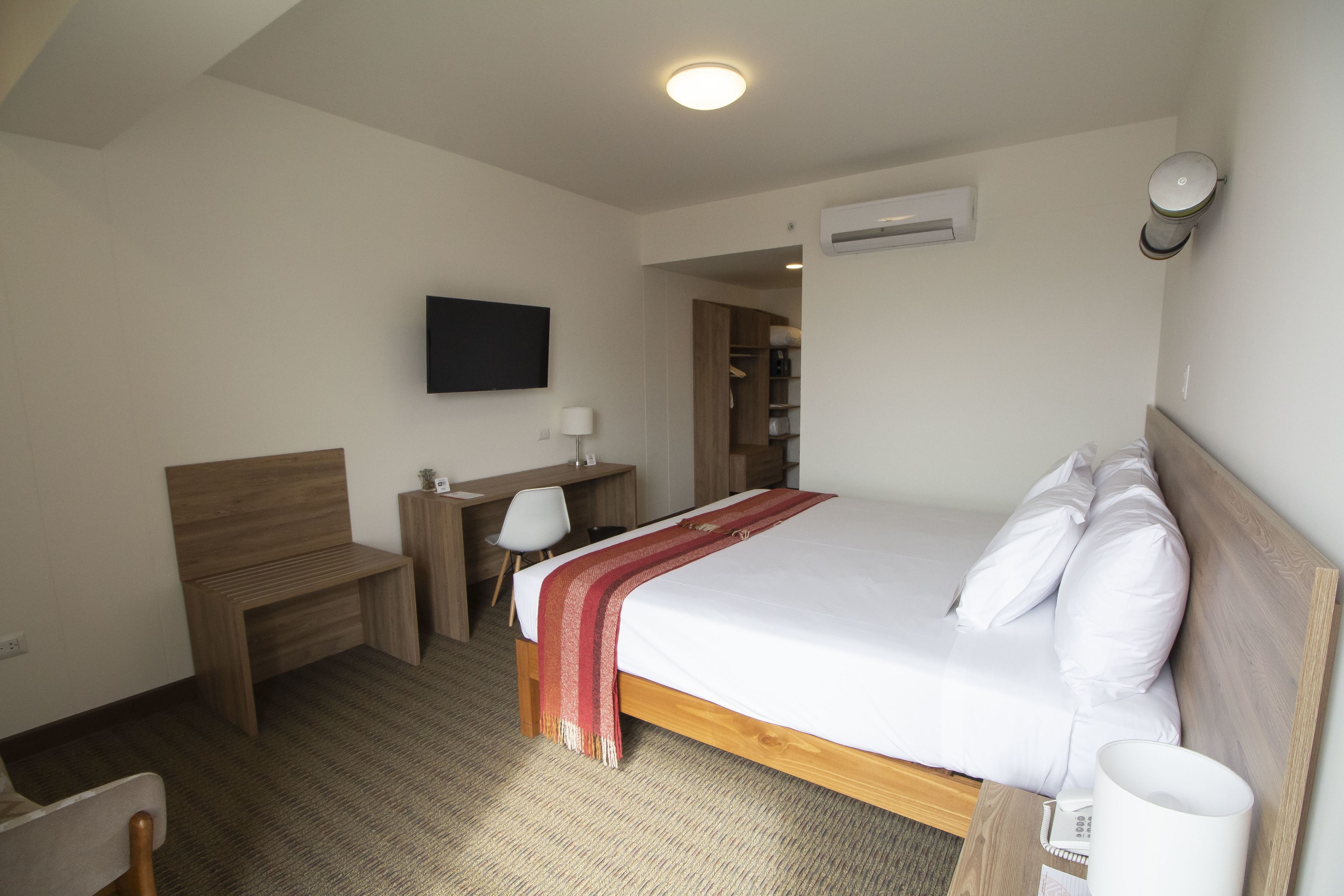 Ein Hotelzimmer mit einem hölzernen Bett, einem Schreibtisch, einem Stuhl, einem an der Wand befestigten Fernseher und einer Klimaanlage.