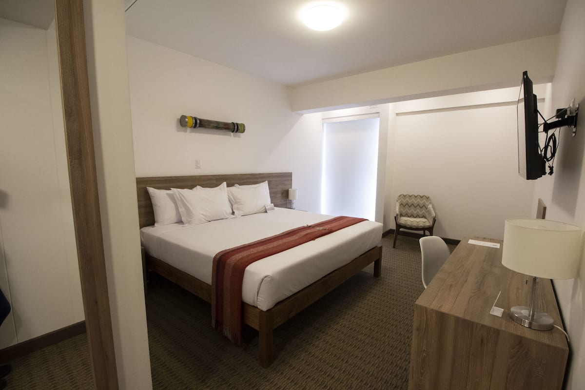 A hotel room with a wooden bed, a desk with a lamp, a chair, and a TV mounted on the wall.