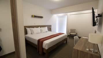 Ein Hotelzimmer mit einem hölzernen Bett, einem Schreibtisch mit Lampe, einem Sessel und einem an der Wand befestigten Fernseher.
