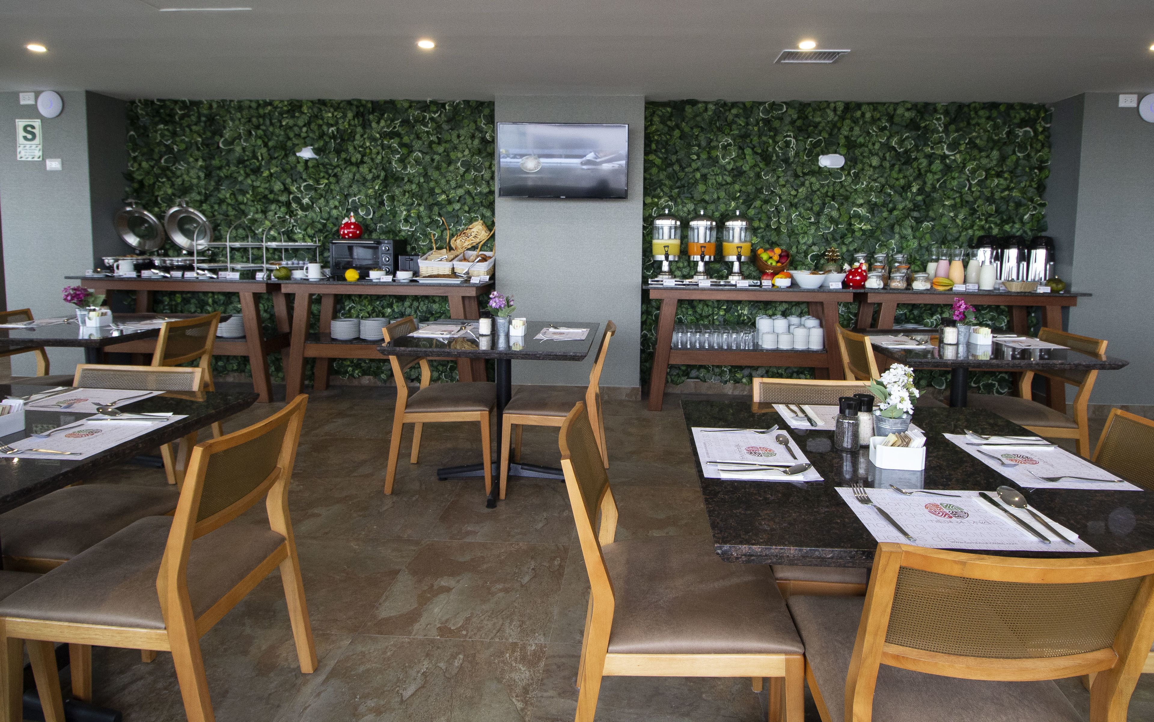 Een restaurant met meerdere tafels en stoelen, een aan de muur bevestigde televisie, en een groene wanddecoratie van bladeren.