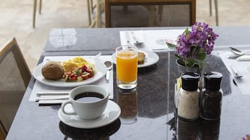 Una mesa de desayuno con huevos revueltos, tostadas y un vaso de jugo de naranja. Hay saleros y pimenteros, una taza de café y un pequeño florero con flores.