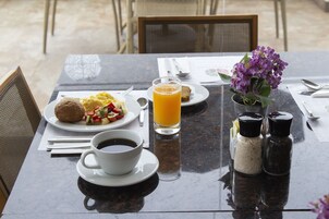Una mesa de desayuno con huevos revueltos, tostadas y un vaso de jugo de naranja. Hay saleros y pimenteros, una taza de café y un pequeño florero con flores.