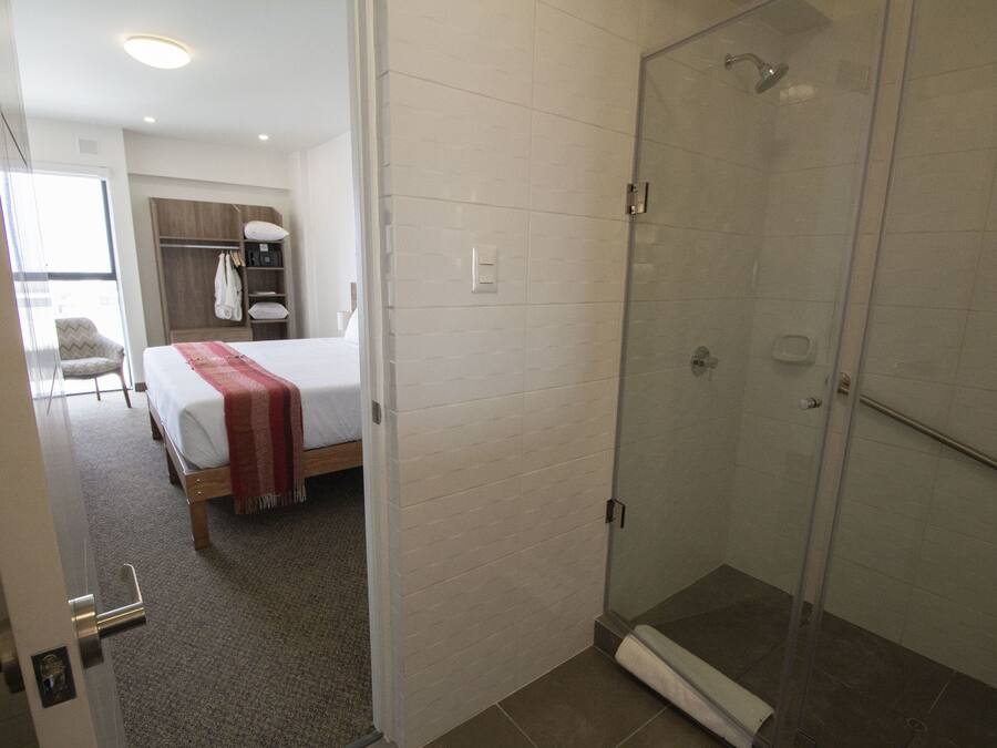Quarto de hotel com cama, closet amplo e área de chuveiro com paredes de vidro.